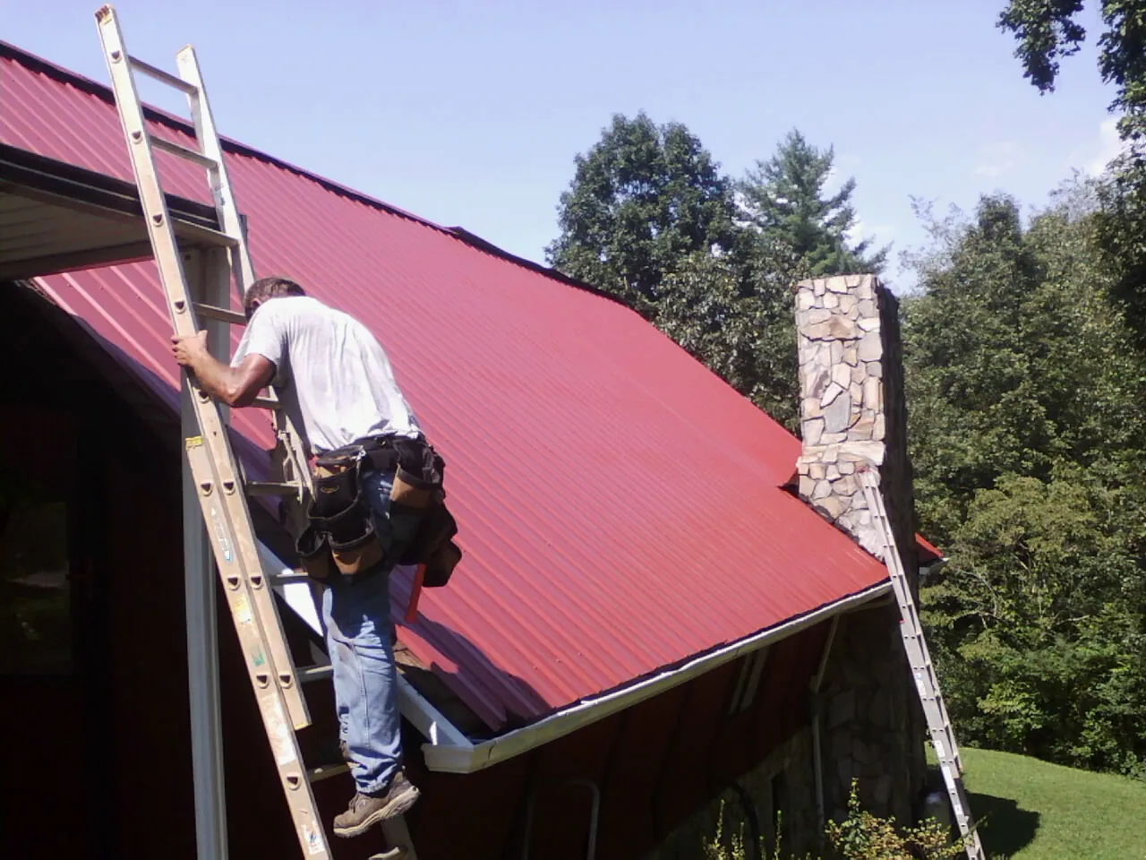 Roofer on a ladder beside a red metal roof and stone chimney — Soffit & Fascia Repair by Roofing Brothers in Lonaconing